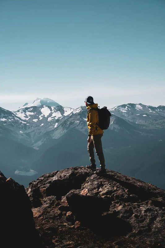 Hiker in yellow jacket on peak with snow-capped mountains; outdoor adventure gear essentials.